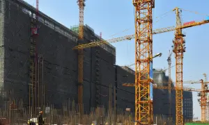Ogromna budowa z wieloma dźwigami, które niczym stalowe szekle spinają niebo. W tle widać budynek otoczony rusztowaniami.