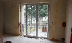 Niedokończona ściana z oknem balkonowym, widok na drzewa i ogrodzenie.