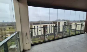 Zabudowa balkonu w bloku z widokiem na nowoczesne budynki. Szklane panele tworzą przytulną przestrzeń.