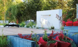 Nowoczesny ogród z betonowymi murkami, drewnianym tarasem i kaskadą wodną. Czerwone donice z roślinami dodają koloru.