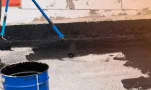 Osoba wykonuje hydroizolację piwnicy od wewnątrz, malując ścianę czarną masą bitumiczną za pomocą wałka.
