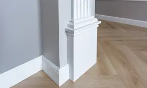 Biały, ozdobny pilaster z żłobieniami, stojący przy szarej ścianie i drewnianej podłodze w jodełkę.