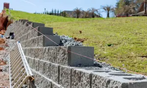 Budowa muru oporowego z bloczków betonowych, z widoczną drabiną i sznurkiem do poziomowania.