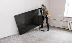 Mężczyzna z pasem narzędziowym przy ścianie trzyma duży, czarny telewizor, zastanawiając się, na jakiej wysokości telewizor będzie najlepiej wyglądał.