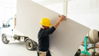 Budowlaniec w żółtym kasku niesie dużą płytę, która posłuży do budowy ściany karton gips.