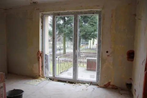 Niedokończona ściana z oknem balkonowym, widok na drzewa i ogrodzenie.