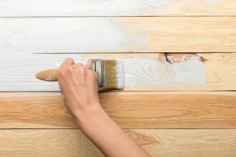 Ręka maluje białą farbą drewnianą boazerię. Proces odnawiania wnętrza trwa.