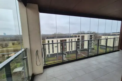 Zabudowa balkonu w bloku z widokiem na nowoczesne budynki. Szklane panele tworzą przytulną przestrzeń.