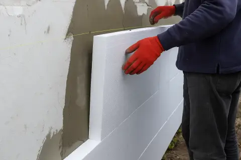 Pracownik w czerwonych rękawicach montuje płyty styropianowe do ściany, rozpoczynając ocieplanie elewacji.