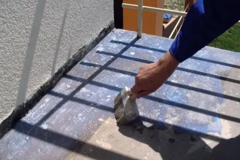 Prace przy remoncie balkonu: ręka z pędzlem nakłada preparat na betonową posadzkę, przygotowując ją do dalszych prac.