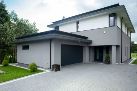 Nowoczesny dom kostka z szarą elewacją, dwupoziomowy, z garażem i podjazdem z kostki brukowej.