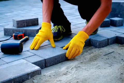 Ręce w żółtych rękawiczkach układają kostkę brukową. Zastanawiasz się, ile kosztuje ułożenie kostki brukowej? Narzędzia takie jak młotek i miarka leżą obok.