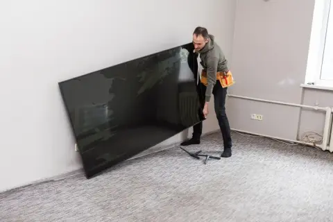 Mężczyzna z pasem narzędziowym przy ścianie trzyma duży, czarny telewizor, zastanawiając się, na jakiej wysokości telewizor będzie najlepiej wyglądał.