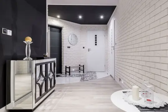 Nowoczesny przedpokój z białymi, ceglanymi ścianami, które nadają mu charakteru. Lustrzana komoda i jasne drewno dodają elegancji.