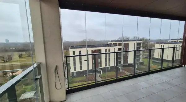 Zabudowa balkonu w bloku z widokiem na nowoczesne budynki. Szklane panele tworzą przytulną przestrzeń.
