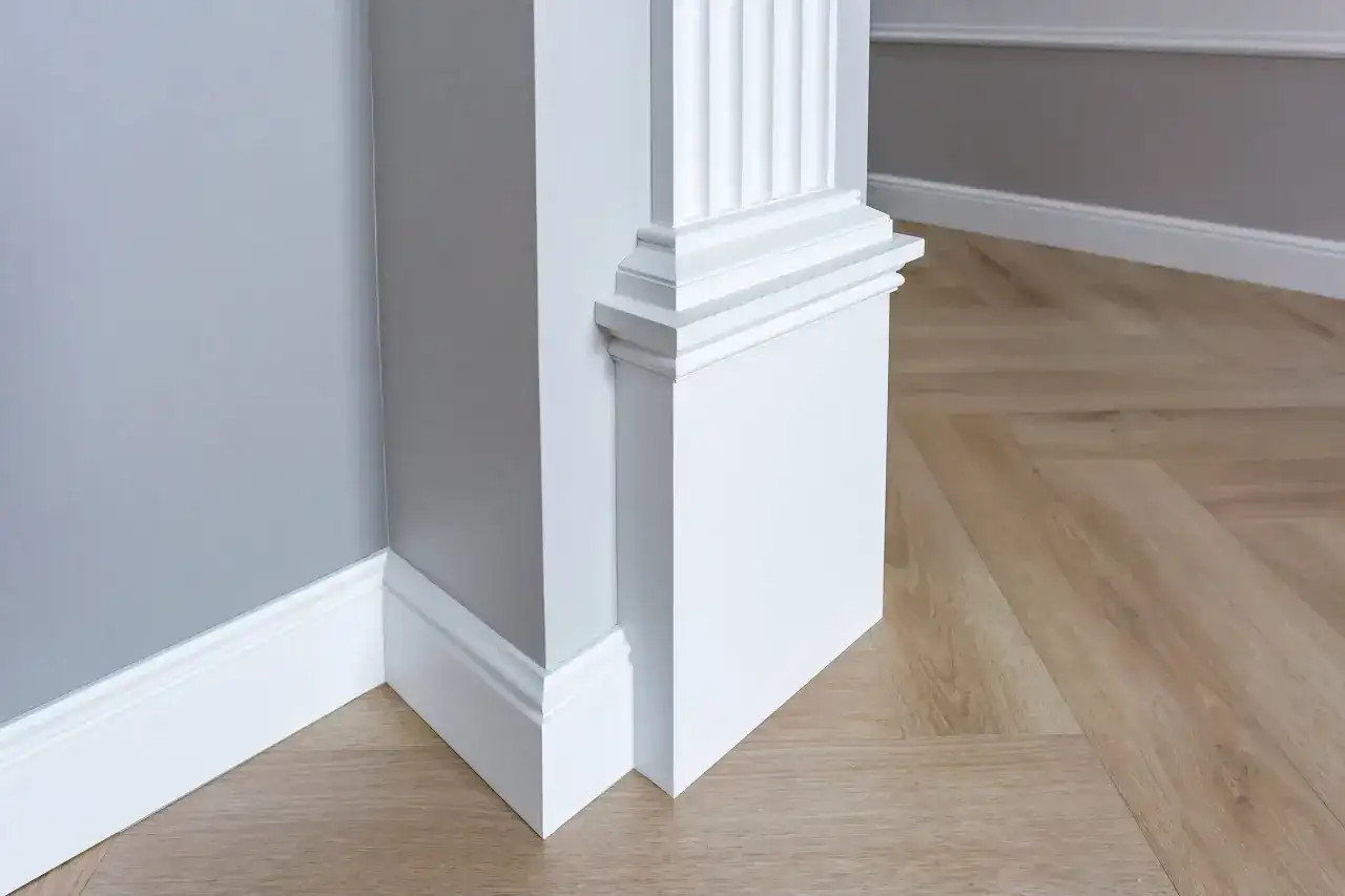 Biały, ozdobny pilaster z żłobieniami, stojący przy szarej ścianie i drewnianej podłodze w jodełkę.