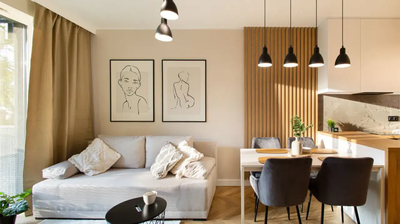 Jak urządzić mały salon z aneksem: jasna sofa, grafiki, drewniana ściana i nowoczesna kuchnia tworzą przytulną przestrzeń.