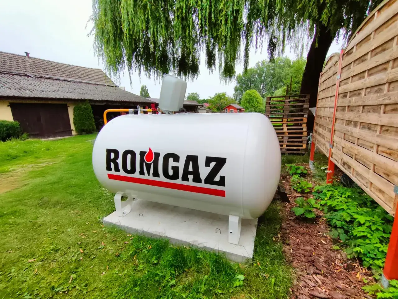 Biały zbiornik na gaz propan z napisem ROMGAZ. W tle dom i drewniany płot.