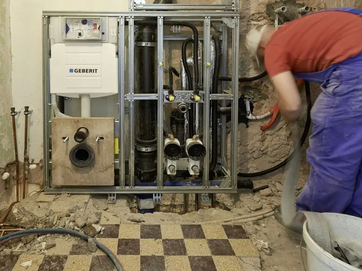 Instalacja Geberit w trakcie prac. Biały montaż, widoczny stelaż, spłuczka i rury. Pracownik w niebieskich ogrodniczkach sprząta gruzy.