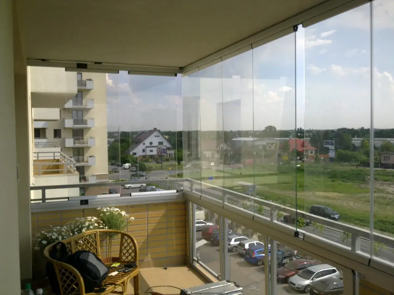 Zabudowa balkonu w bloku z widokiem na ulicę i domy. Na balkonie rattanowe krzesło z plecakiem i kwiatami.