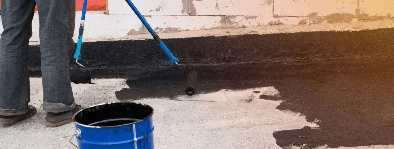 Osoba wykonuje hydroizolację piwnicy od wewnątrz, malując ścianę czarną masą bitumiczną za pomocą wałka. Obok stoi niebieska beczka z materiałem.