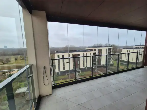 Zabudowa balkonu w bloku z widokiem na nowoczesne budynki. Szklane panele tworzą przytulną przestrzeń.