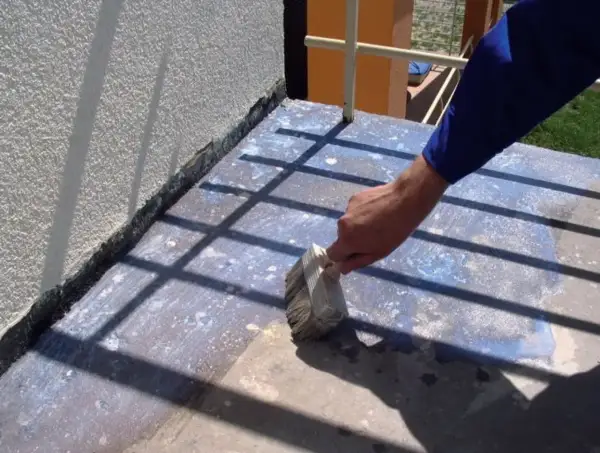 Prace przy remoncie balkonu: ręka z pędzlem nakłada preparat na betonową posadzkę, przygotowując ją do dalszych prac.