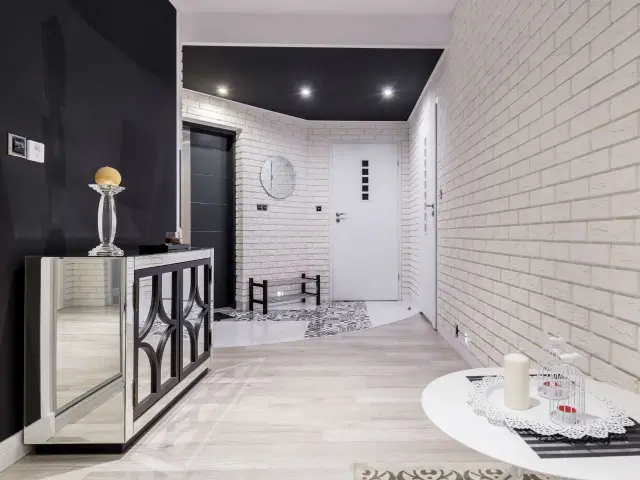 Nowoczesny przedpokój z białymi, ceglanymi ścianami, które nadają mu charakteru. Lustrzana komoda i jasne drewno dodają elegancji.