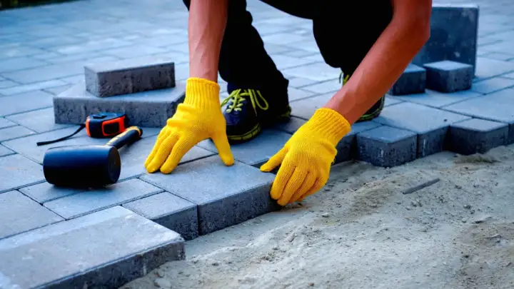Ręce w żółtych rękawiczkach układają kostkę brukową. Zastanawiasz się, ile kosztuje ułożenie kostki brukowej? Narzędzia takie jak młotek i miarka leżą obok.